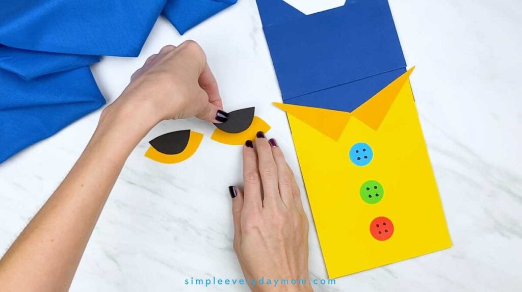 Hands layer a black paper eye shape onto a yellow eye piece beside the partially assembled Pete the Cat puppet.