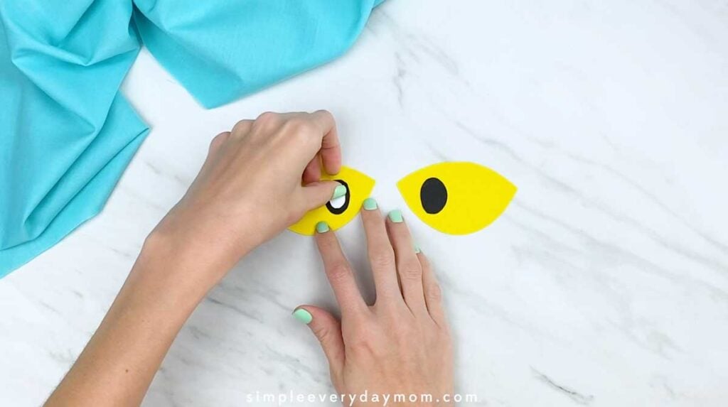 Hands adding a white highlight to the black pupil on a yellow paper eye.