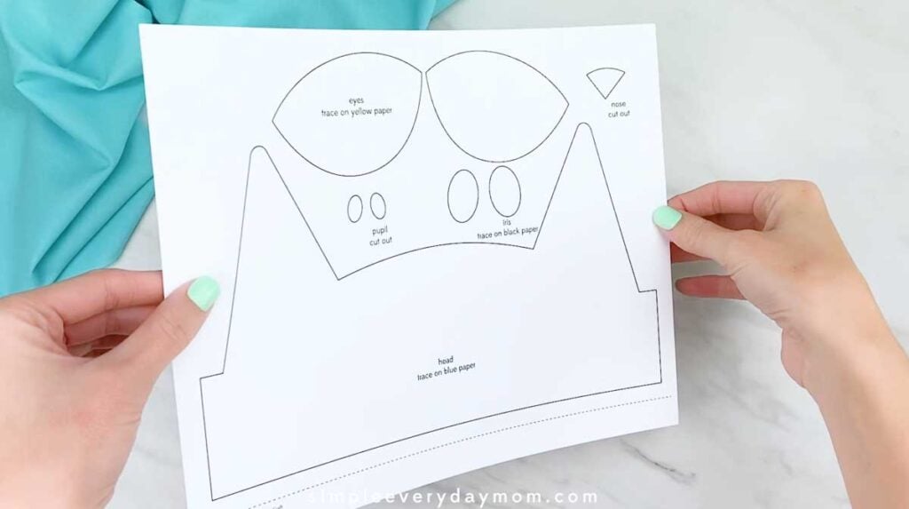 Hands holding a printable template for a blue cat headband with eye, pupil, nose, and head pieces.