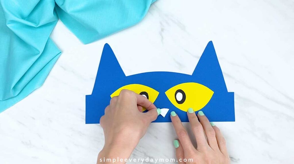 Hands attaching a white triangular nose between the cat’s yellow eyes.