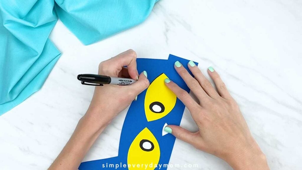 Hand outlining a yellow eye with black marker on the blue cat headband.