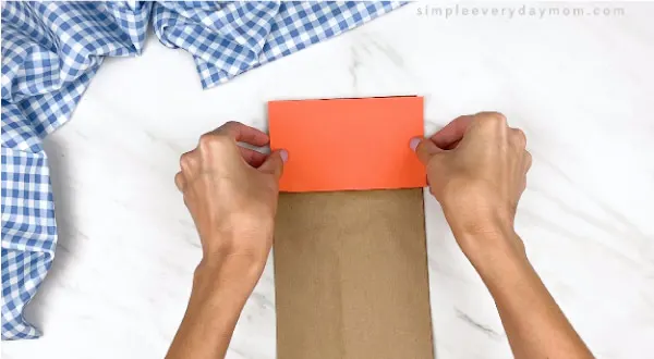 hands gluing on orange paper to top of brown paper bag