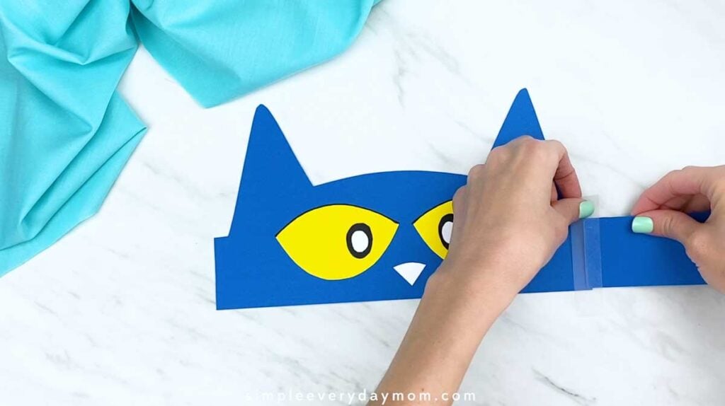Hands taping a blue paper strap to one side of the cat headband.