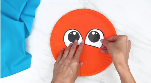 hands gluing on paper eyes onto orange paper plate 