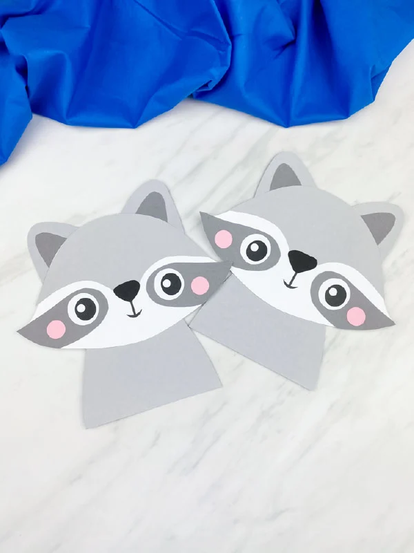two completed paper raccoon crafts on marble background with blue fabric 