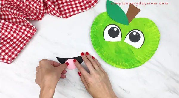 hands gluing paper tongue on paper mouth, with paper plate apple to the right 