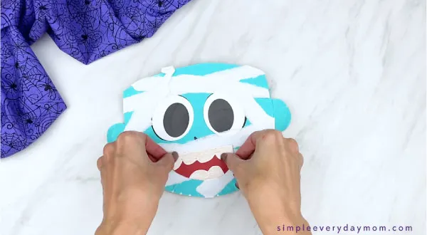 hands gluing mouth onto blue paper plate mummy craft