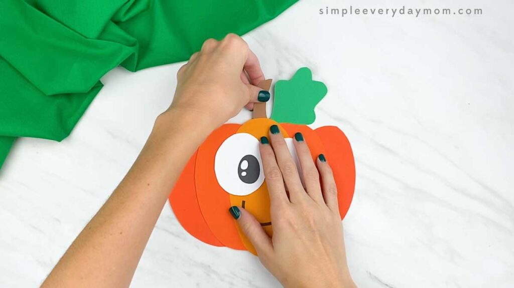 Hands glue a brown paper stem behind the pumpkin leaf.