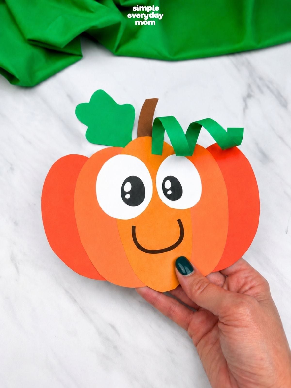 Close-up of a hand holding a completed smiling paper pumpkin craft with layered orange paper, large cartoon eyes, a green leaf, curled vine, and brown stem.