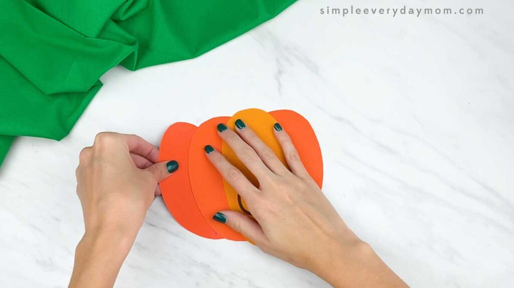 Hands attach another orange pumpkin section to build the layered paper pumpkin shape.