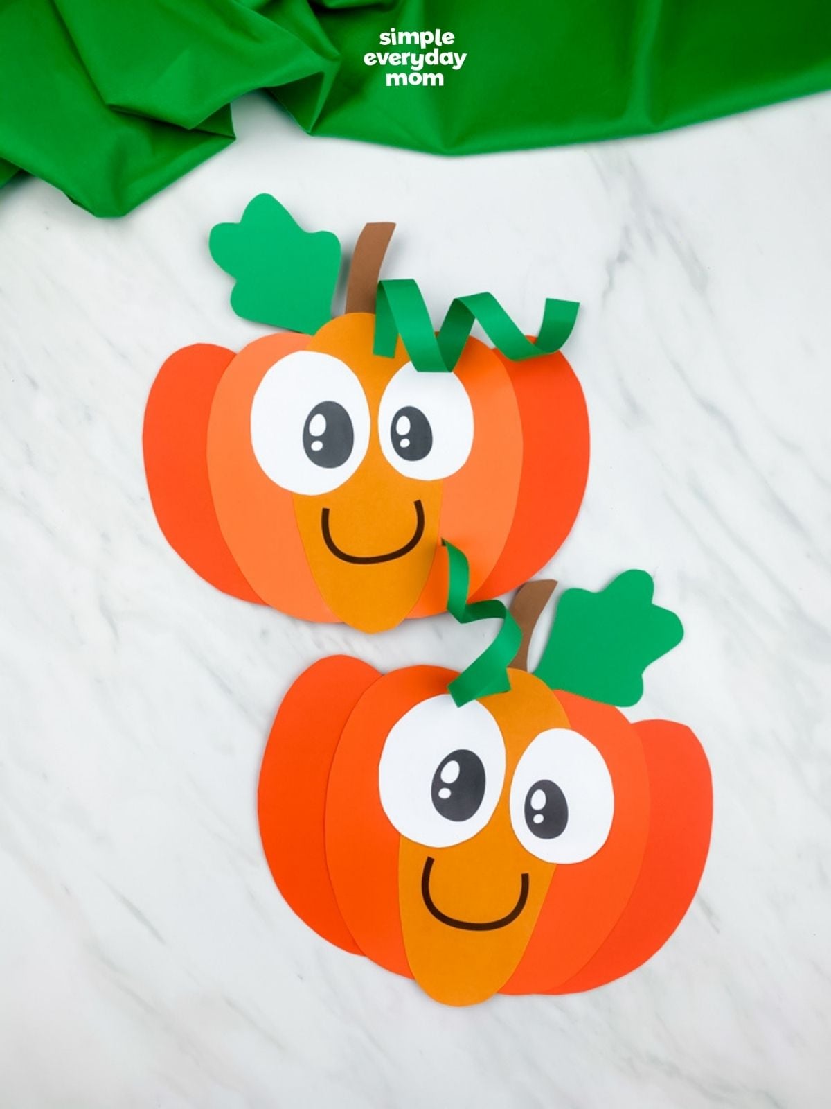 Two completed smiling paper pumpkin crafts are arranged vertically on a white marble background.