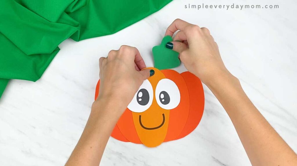 Hands attach a green paper leaf to the top of the completed pumpkin craft.