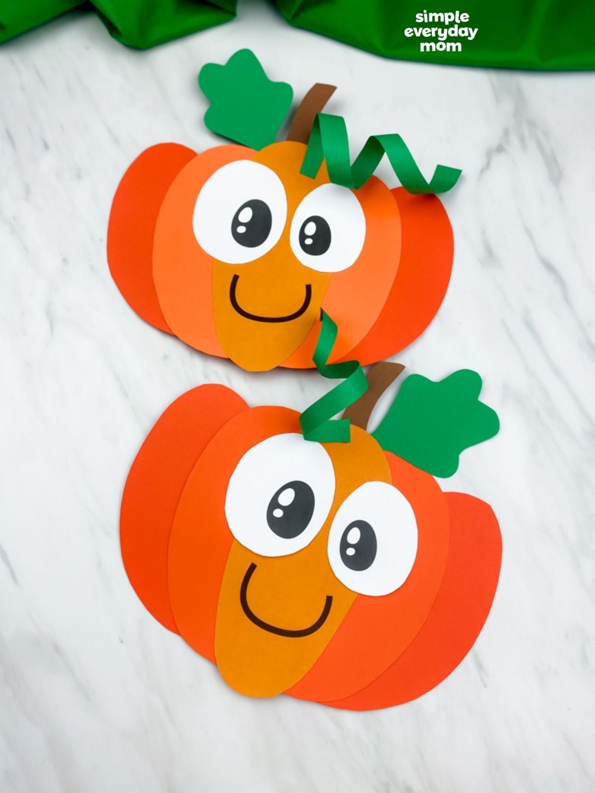 Two completed smiling paper pumpkin crafts with curled green vines are displayed on a white marble surface.