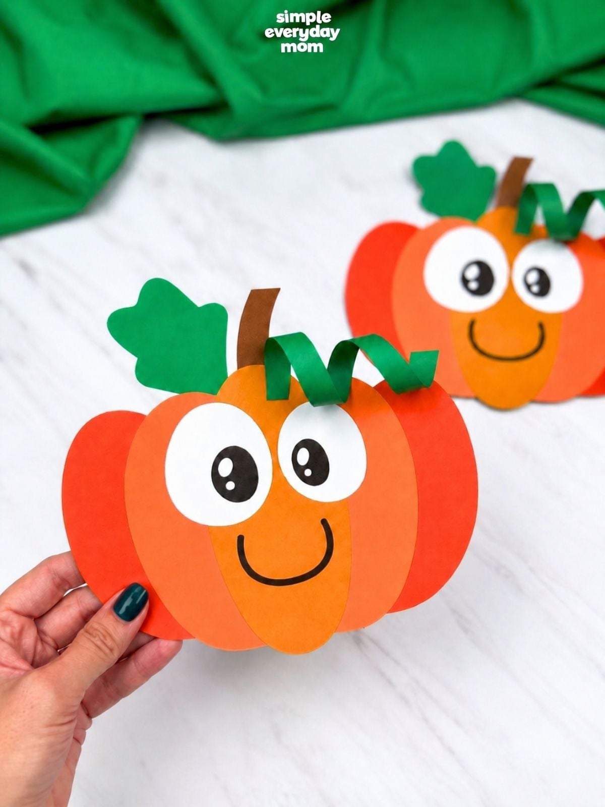 Close-up of a smiling paper pumpkin craft in the foreground with a second finished pumpkin blurred in the background.