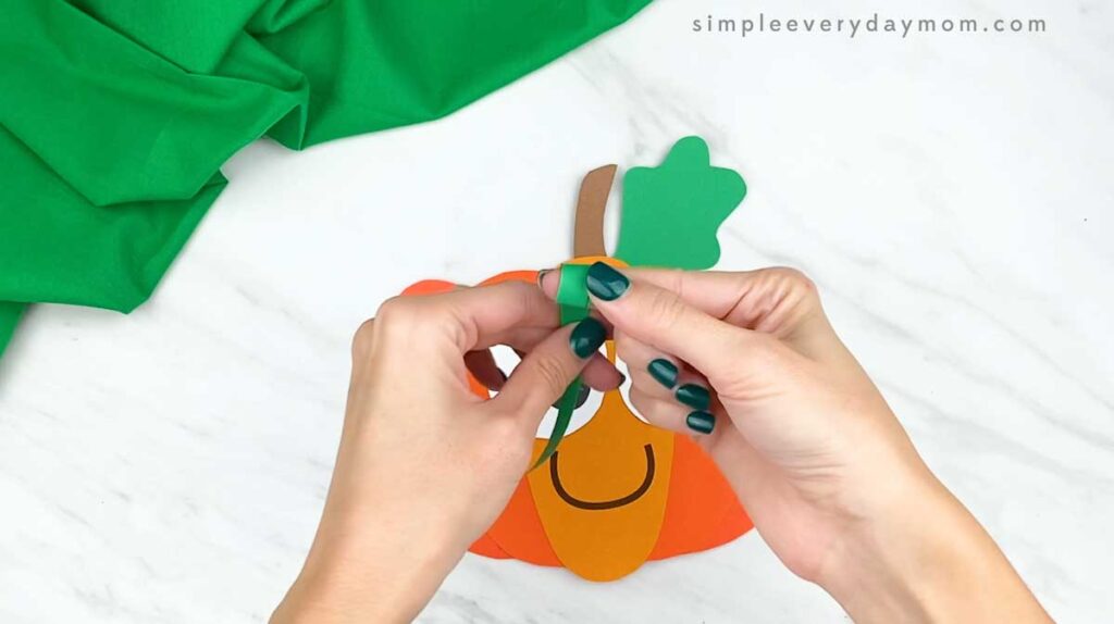 Hands curl a strip of green paper to create a spiral pumpkin vine.