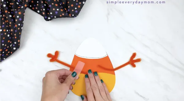 hands taping pipe cleaner arms to the back of paper candy corn craft