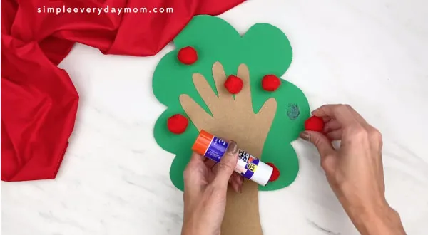 hands gluing red pom poms onto paper tree