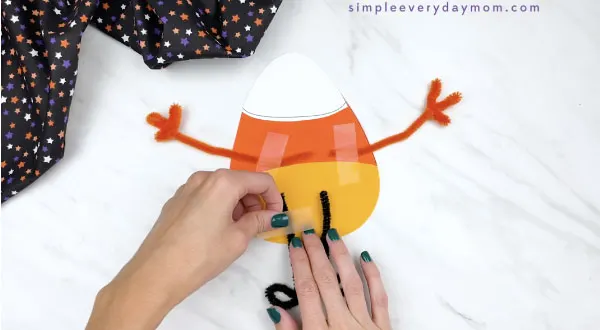 hands taping pipe cleaner legs to the back of paper candy corn craft