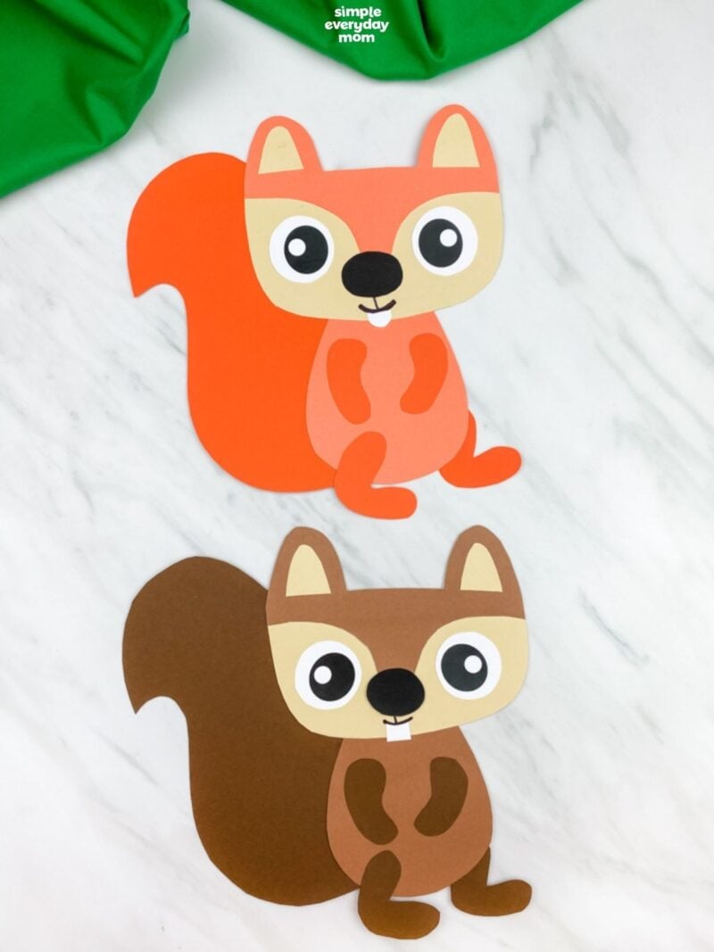 Two paper squirrel crafts, one orange and one brown, are displayed on a white marble surface beneath green fabric.