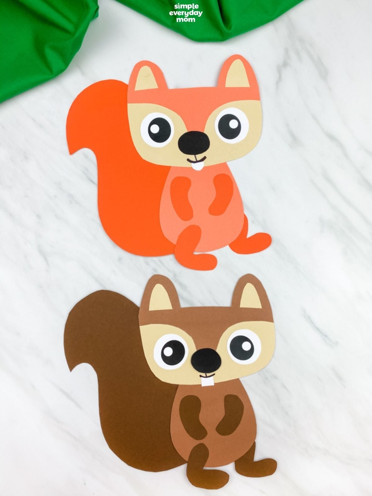 Two paper squirrel crafts, one orange and one brown, are displayed on a white marble surface beneath green fabric.