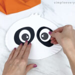 hands gluing eyes to paper plate panda craft