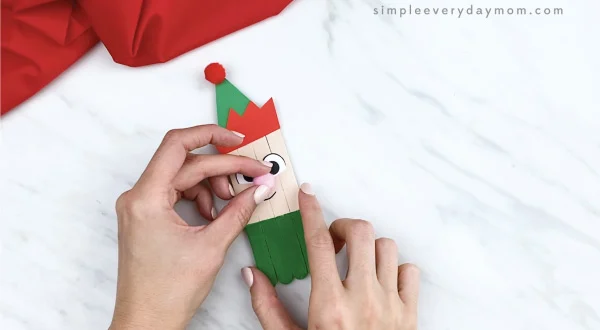 hands gluing pom pom nose onto popsicle stick elf craft