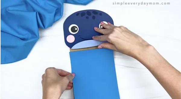 hands gluing blue rectangle to paper bag whale craft