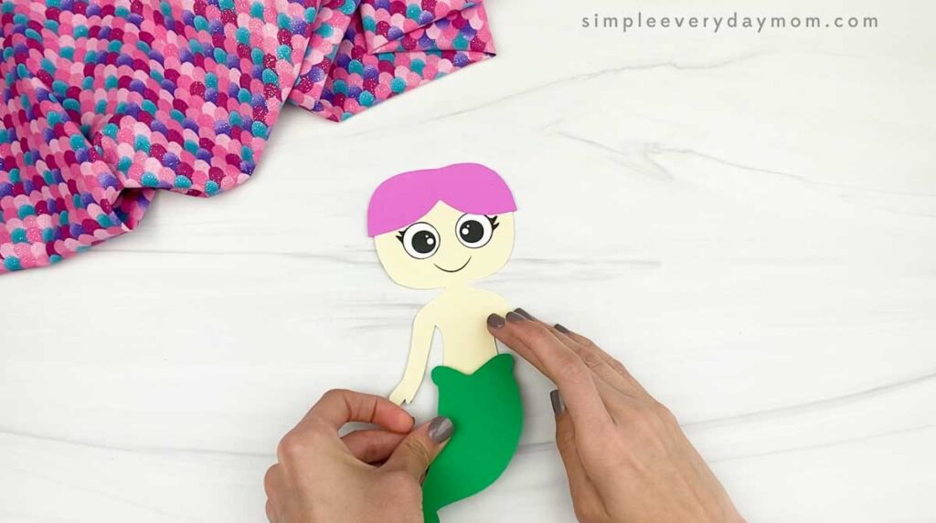Hands attach a green tail piece to a paper mermaid body.