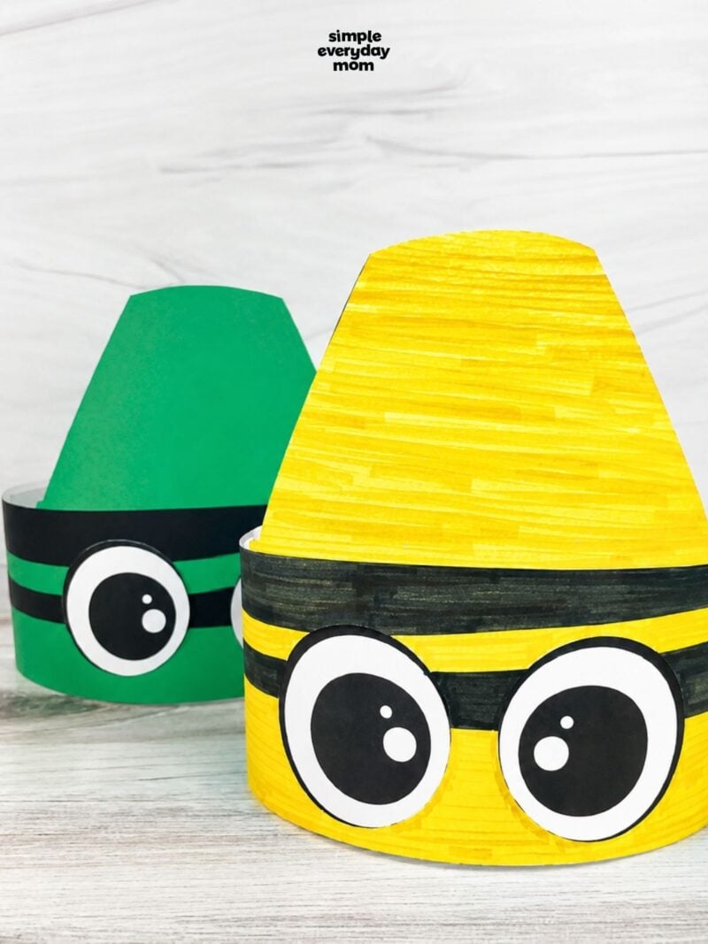Yellow and green crayon-shaped paper headbands with large cartoon eyes displayed on a light wood surface.