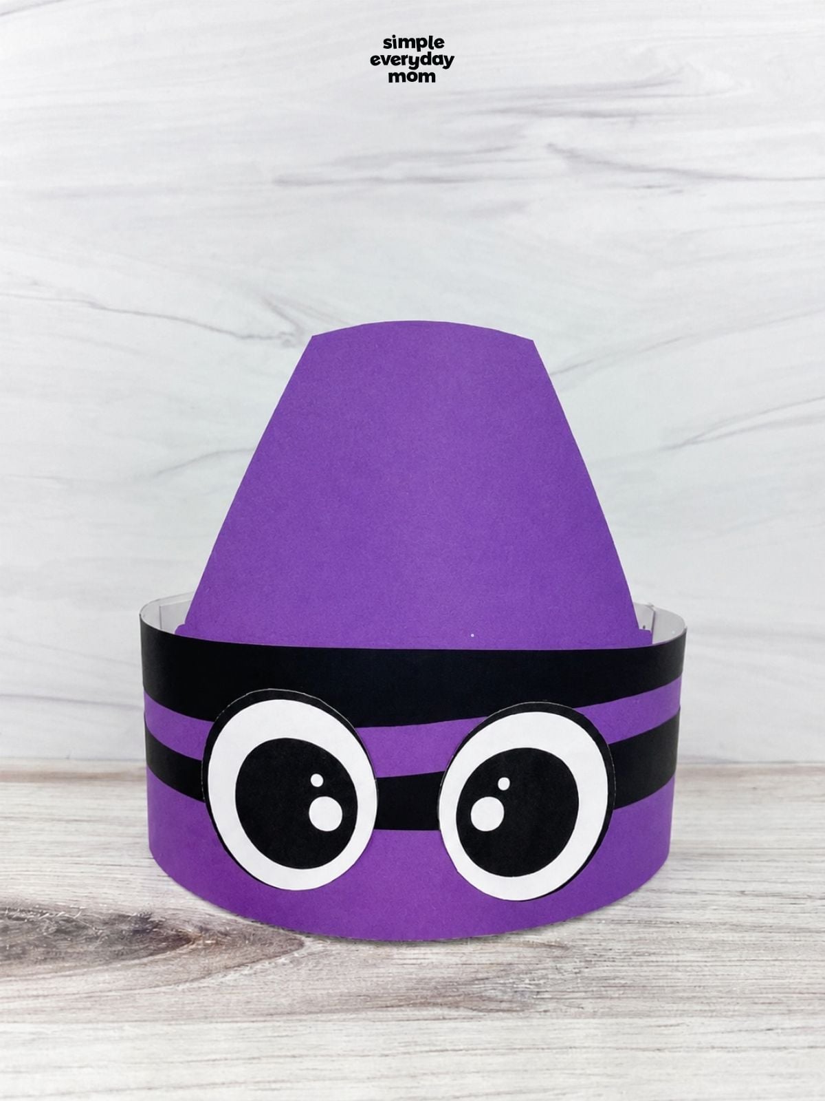 Purple crayon-shaped paper headband with large cartoon eyes displayed on a light wood surface.