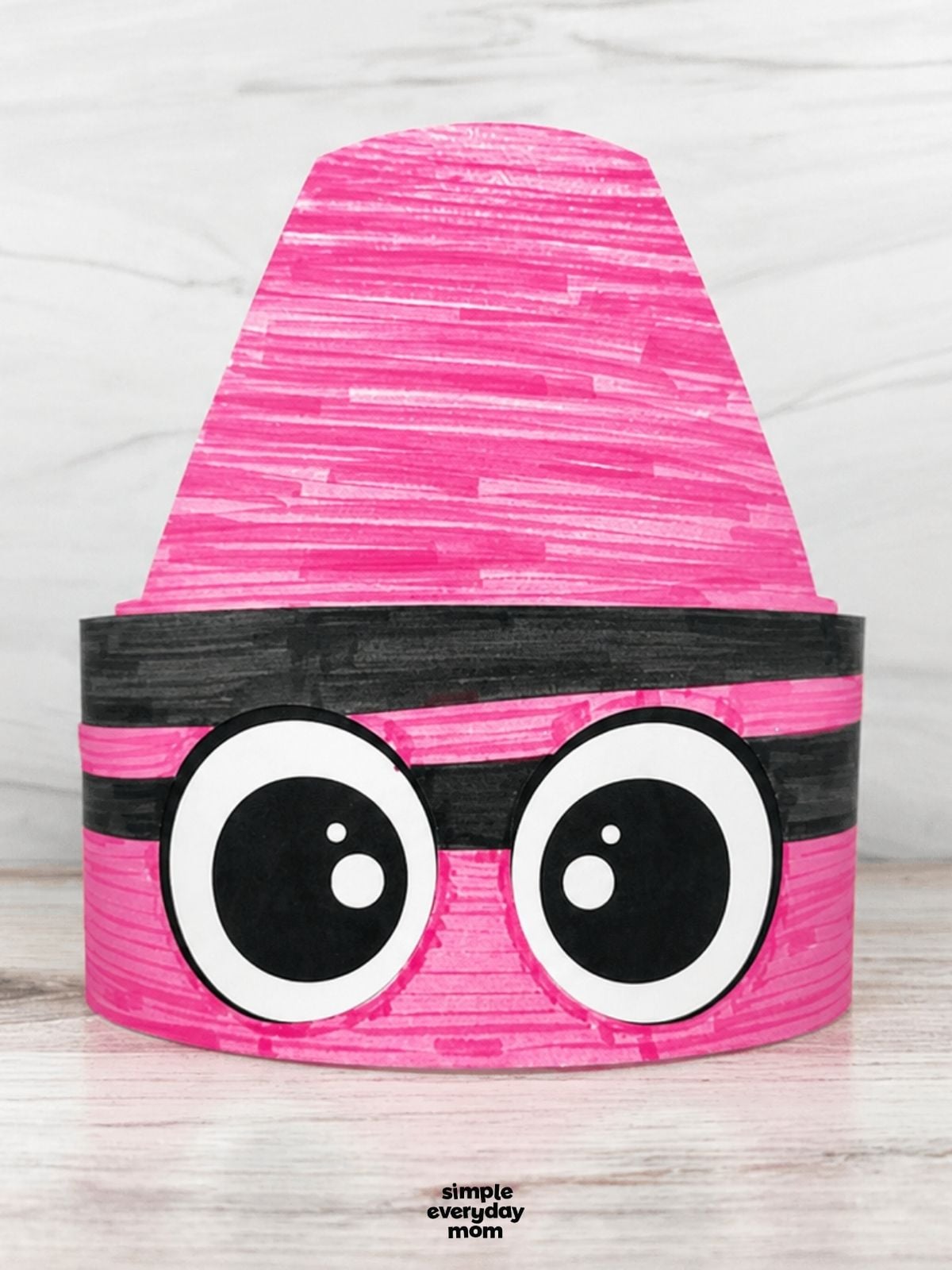 Pink crayon-shaped paper headband with large cartoon eyes displayed on a light wood surface.
