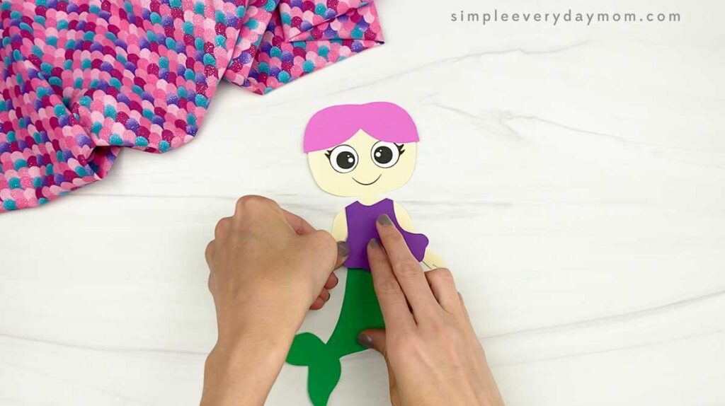 Hands add a purple top to a partially assembled paper mermaid.