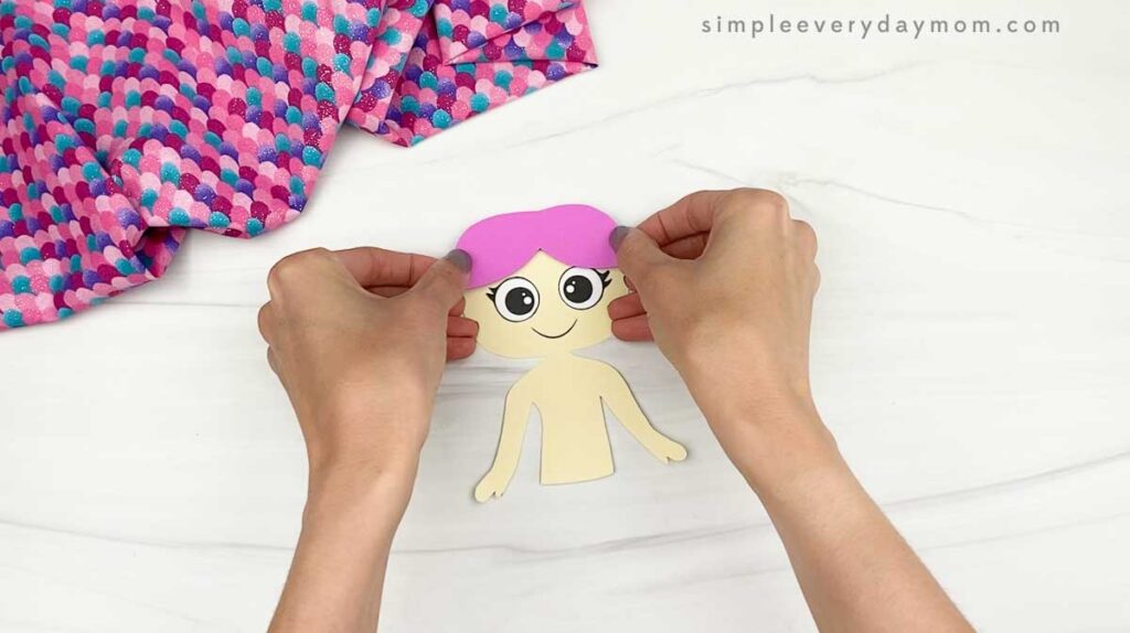 Hands place pink paper hair onto a mermaid face during the craft assembly.