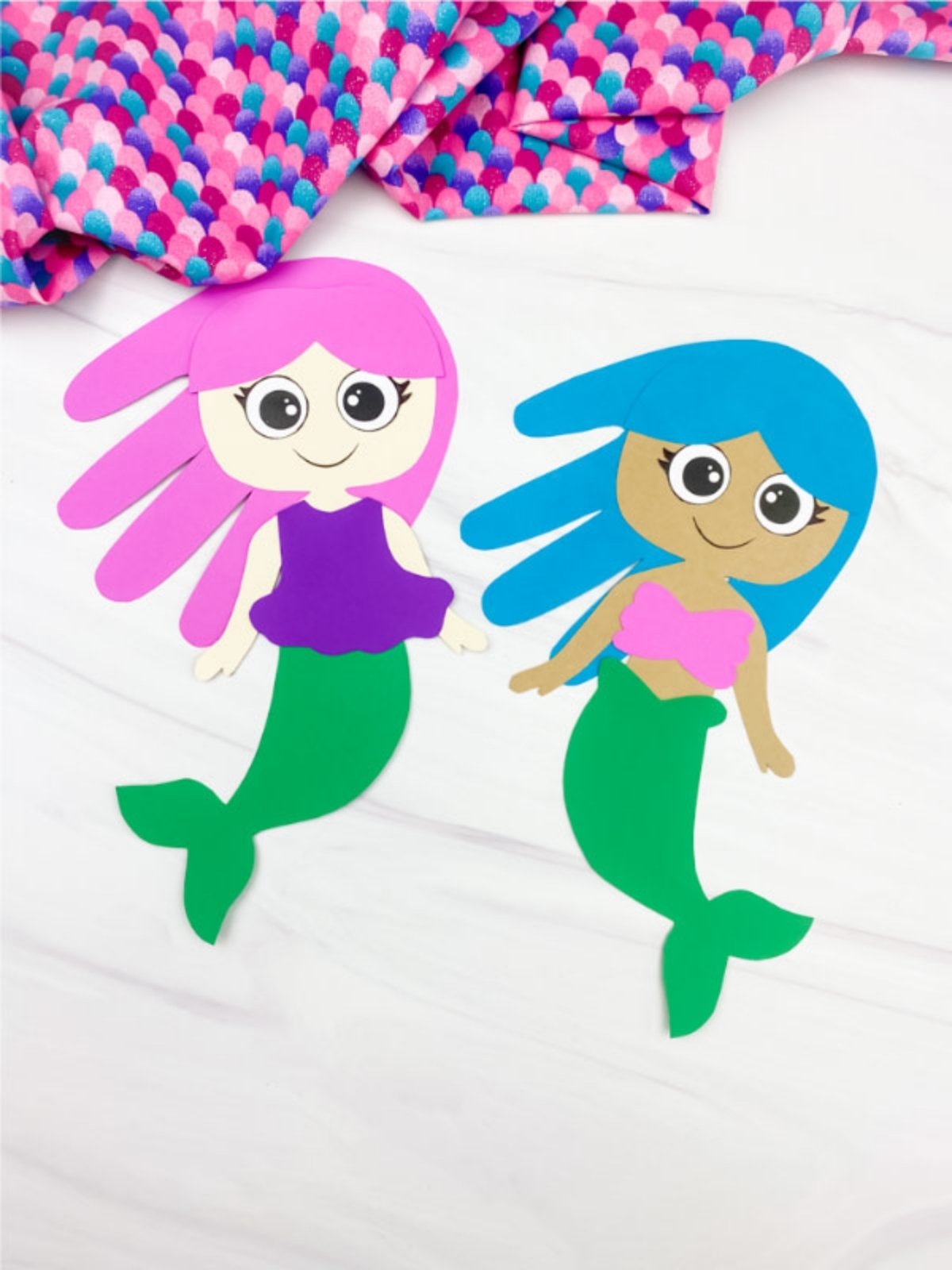 Two finished paper mermaid crafts with colorful hair and green tails displayed on a white surface.
