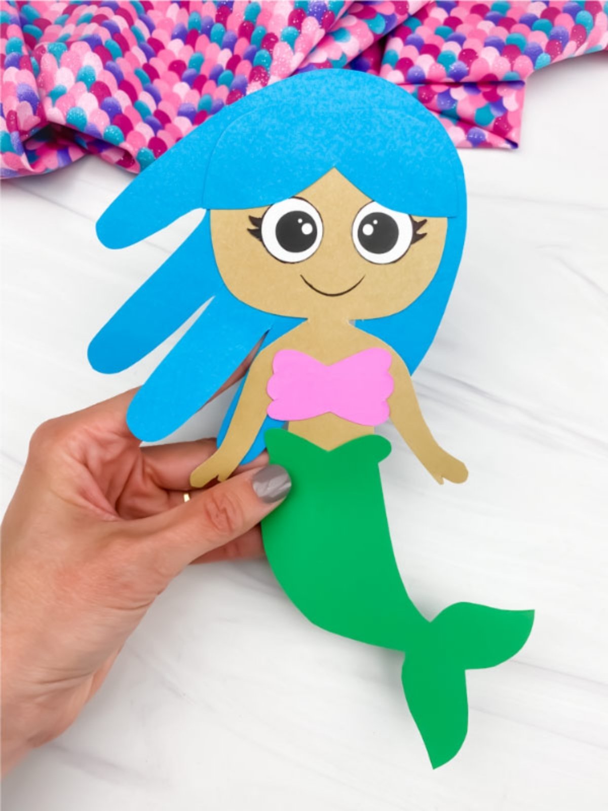 Close-up of a hand holding a completed paper mermaid craft with blue hair and a green tail.