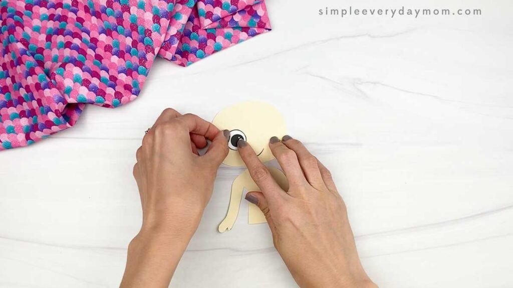 Hands attach eyes to a paper cutout of a mermaid body on a white surface.