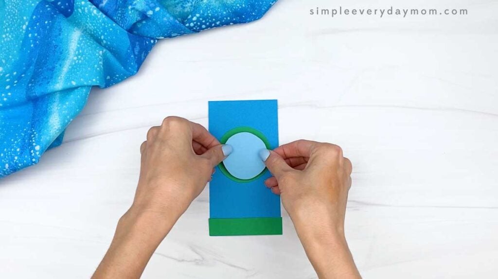 Hands adding a light blue circle inside the green frame to form the rocket window.