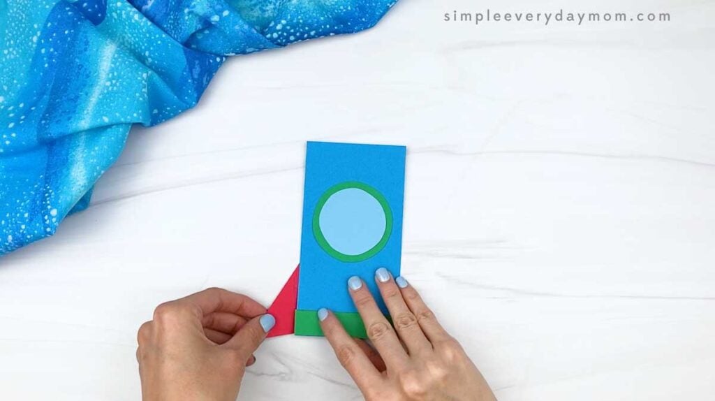 Hands attaching a red triangular fin to the side of the paper rocket.