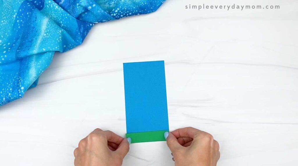 Hands attaching a green strip to the bottom of a blue paper rectangle.