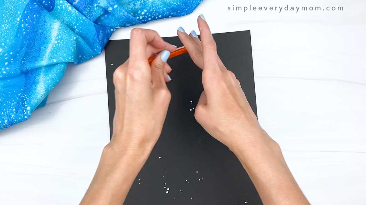 Hands splattering white paint onto black paper to create a starry background.