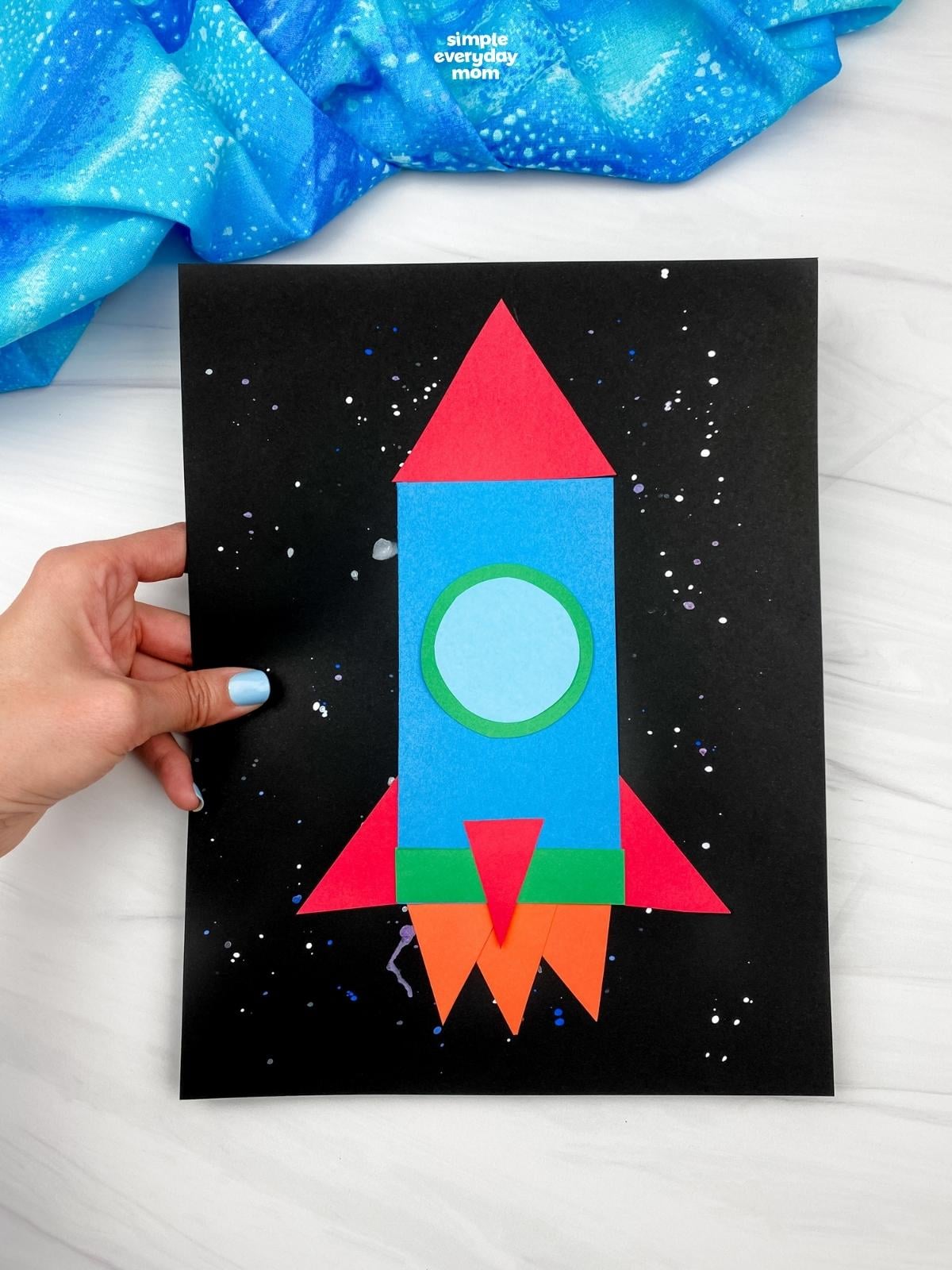 Hand holding a blue paper rocket craft with red fins, orange flames, and a circular window.