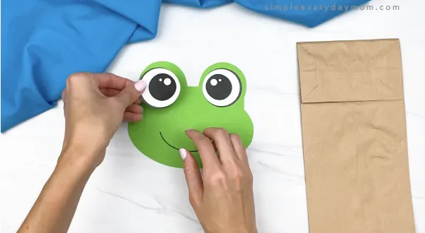 hand gluing eye to paper bag frog head