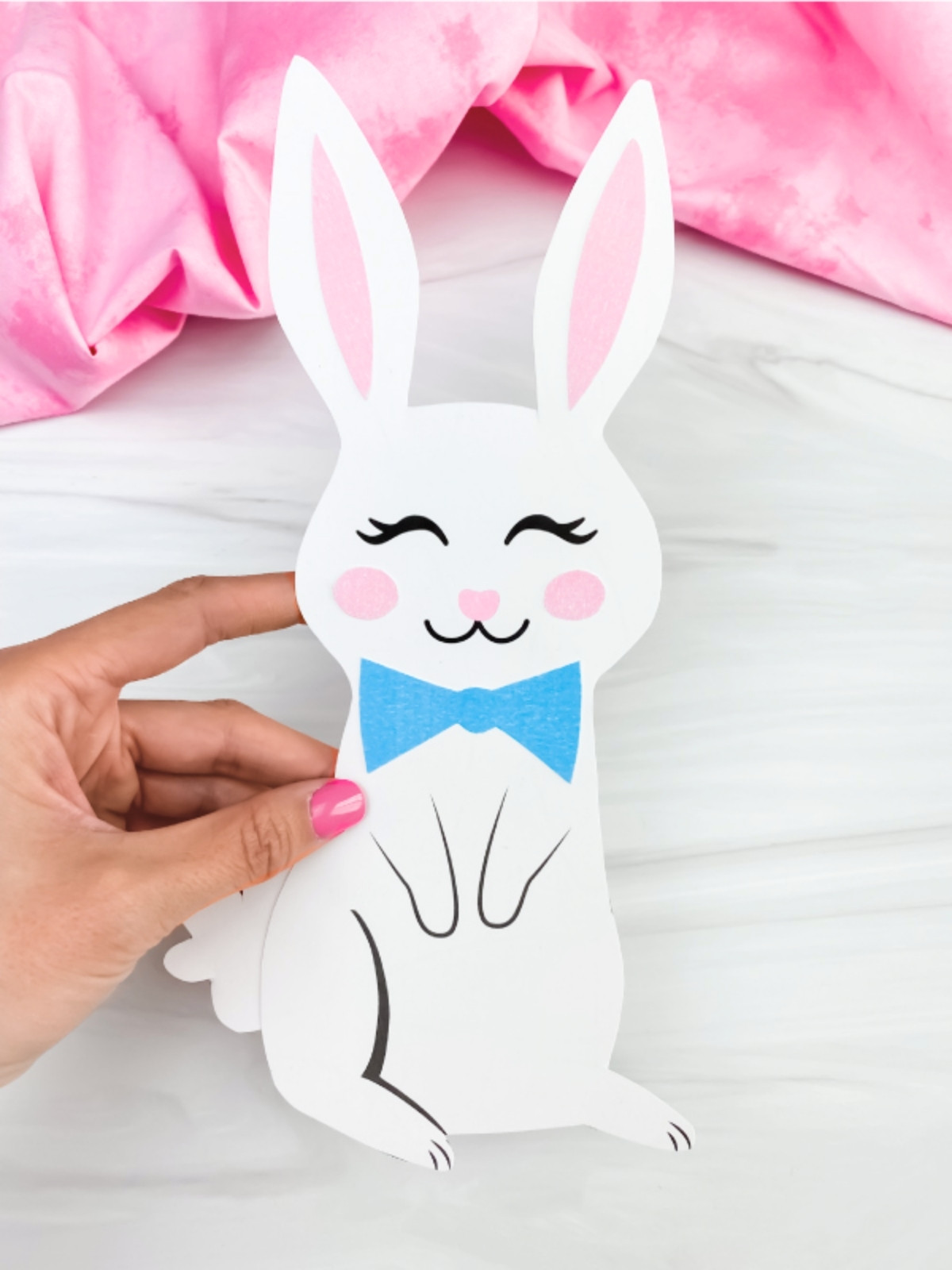 Hand holding a white paper bunny with a blue bow tie on a light surface.