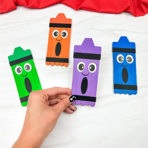 hand holding purple popsicle stick crayon craft with green, red, and blue crafts in the background