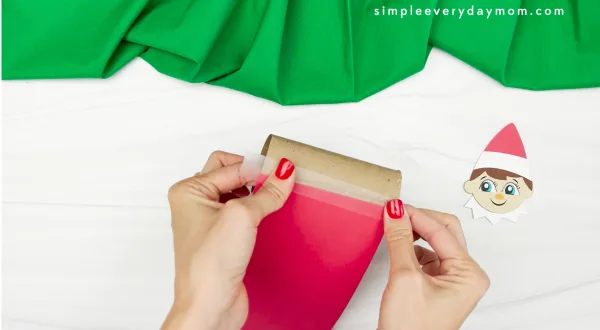hands assembling the body of the elf on the shelf toilet paper roll, taping red paper to the roll