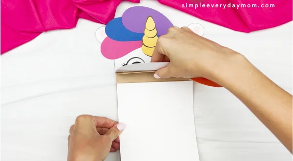 hands gluing body onto paper bag of unicorn rainbow puppet