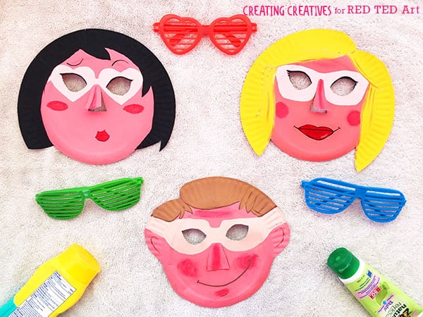 Three decorated paper plate face masks with cut-out eyes are arranged with colorful toy sunglasses and paint tubes.
