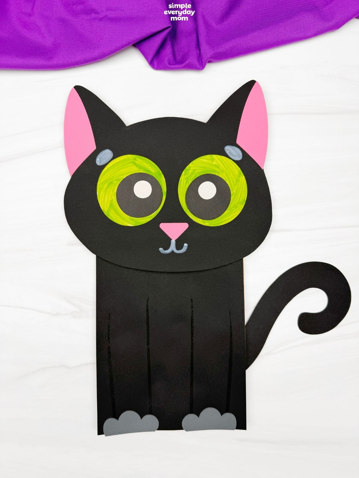 Finished black paper cat puppet with oversized green eyes, pink ears and nose, gray paws, and a curled black tail.