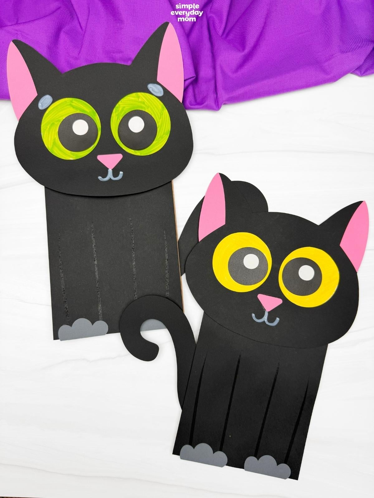 Two finished black paper cat puppets with pink ears, curled tails, gray paws, and oversized green and yellow eyes displayed on a white surface.