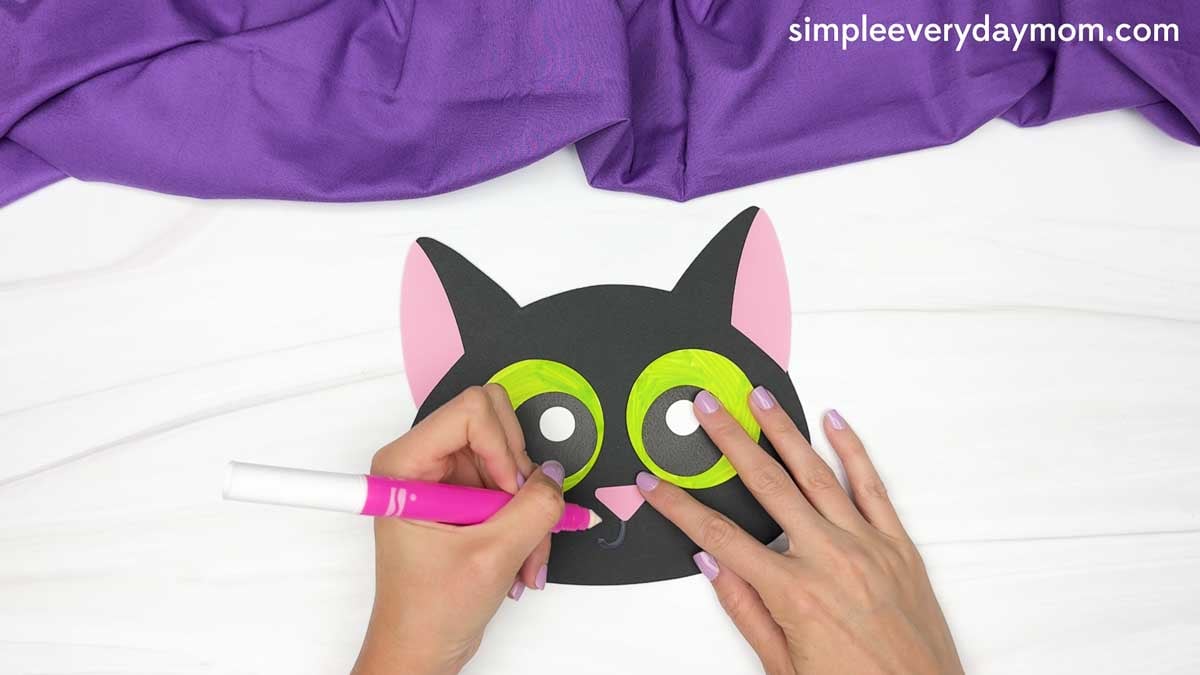 A hand uses a marker to draw the cat’s mouth beneath its pink nose on the black paper face.